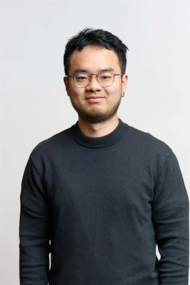 A young East Asian man in glasses and a dark sweater smiles subtly.