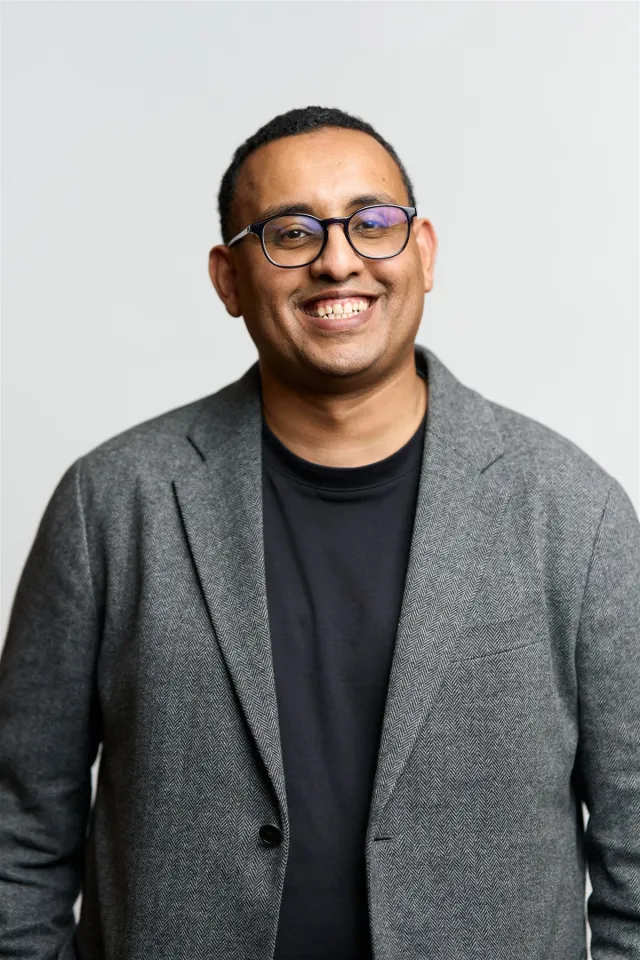 Smiling man in glasses and a grey blazer over a black shirt.